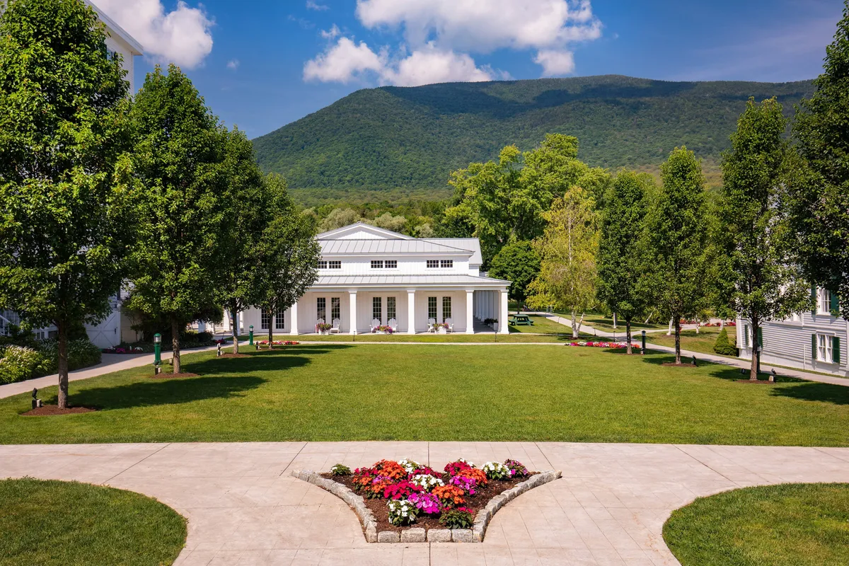 The image features the resort’s grand exterior with stately columns, beautifully landscaped gardens, and a stunning mountain backdrop. The scene captures moments of relaxation in the garden and the expansive, welcoming grounds.