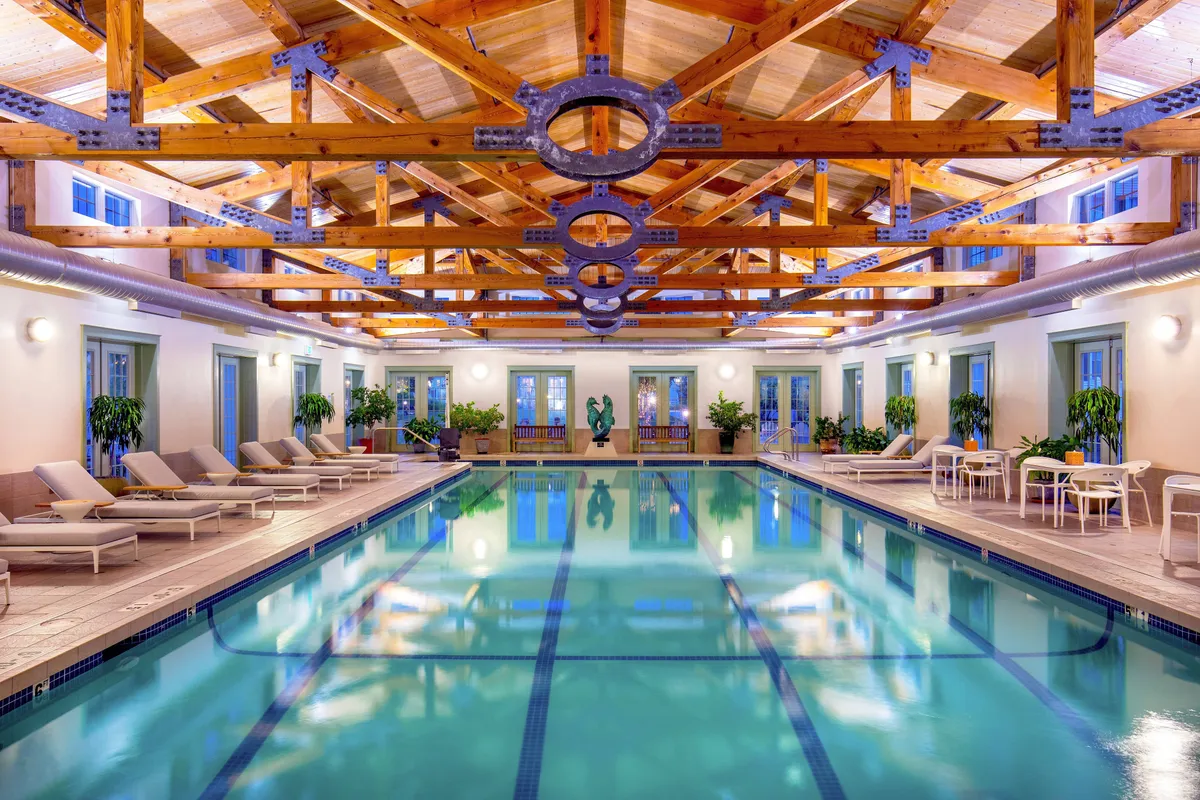 A luxurious indoor swimming pool area with elegant wooden beams above, comfortable lounge chairs, potted plants, and a central sculpture. The lighting and decor create a soothing, upscale ambiance ideal for unwinding.