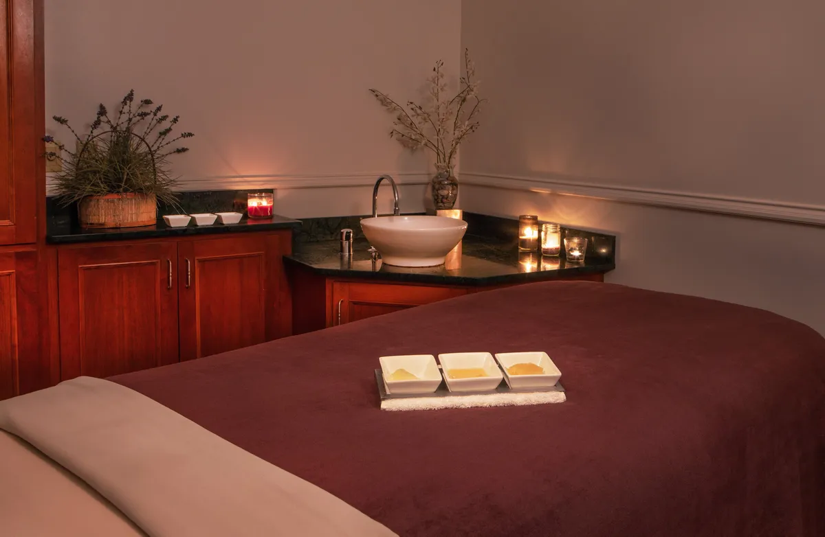 A tranquil spa setting featuring a massage table surrounded by decorative plants, a bowl sink, and softly glowing candles. The space exudes a calming atmosphere, suggestive of personal relaxation and rejuvenation.