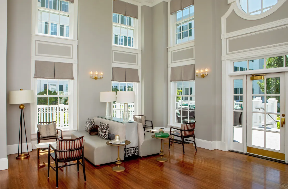 This inviting interior image features a plush sofa adorned with decorative pillows, a cozy coffee table with a candle, floor-to-ceiling windows that flood the room with natural light, a stylish lamp, and an outdoor patio visible through glass doors. The scene radiates comfort and a welcoming atmosphere, hinting at a space where relaxation, socializing, and enjoying the scenic views are effortless.