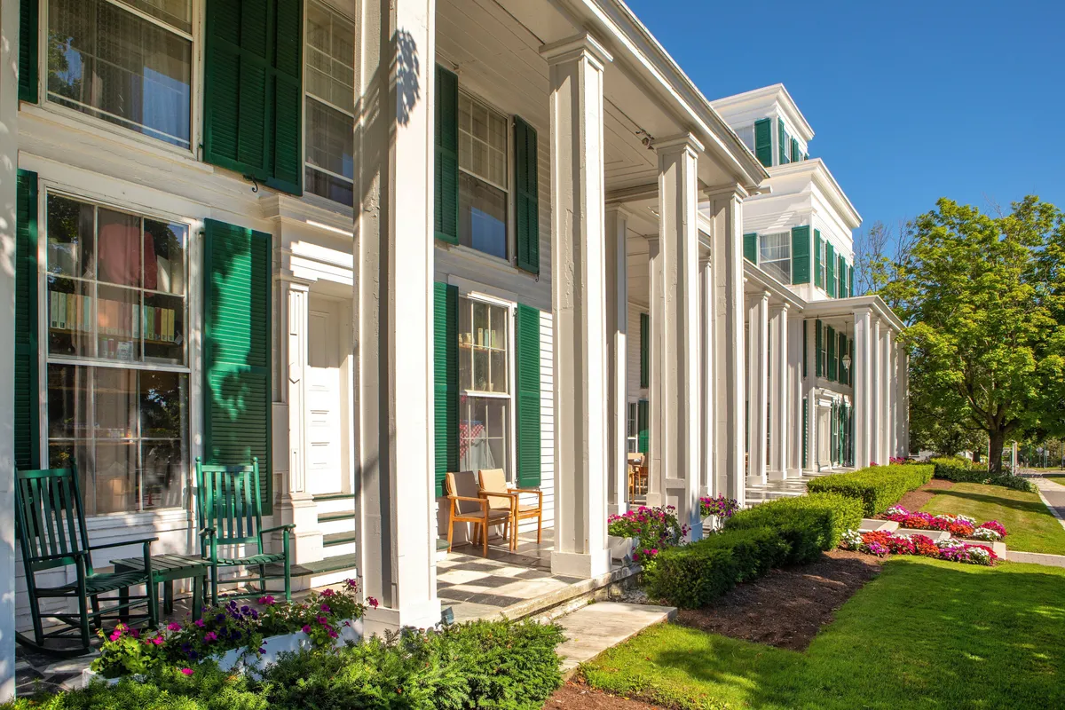 The image showcases the inviting exterior of The Equinox Resort, featuring classic green shutters, white columns, and a welcoming porch lined with rocking chairs and vibrant flower beds. The scene radiates relaxation, with guests enjoying the peaceful outdoor setting—some reading, others simply taking in the natural beauty. The ambiance is both historic and genteel, reflecting the resort's luxurious hospitality and its connection to Vermont's landscape.