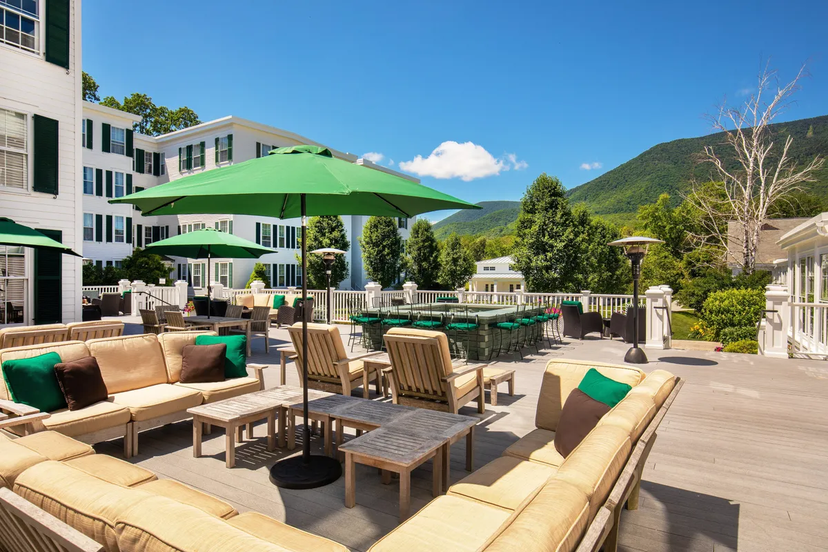 A spacious outdoor dining area set under green umbrellas with tables inviting guests to socialize, dine, and enjoy panoramic mountain views. The casual setting encourages guests to savor Vermont cuisine while basking in the natural beauty of the resort’s grounds.