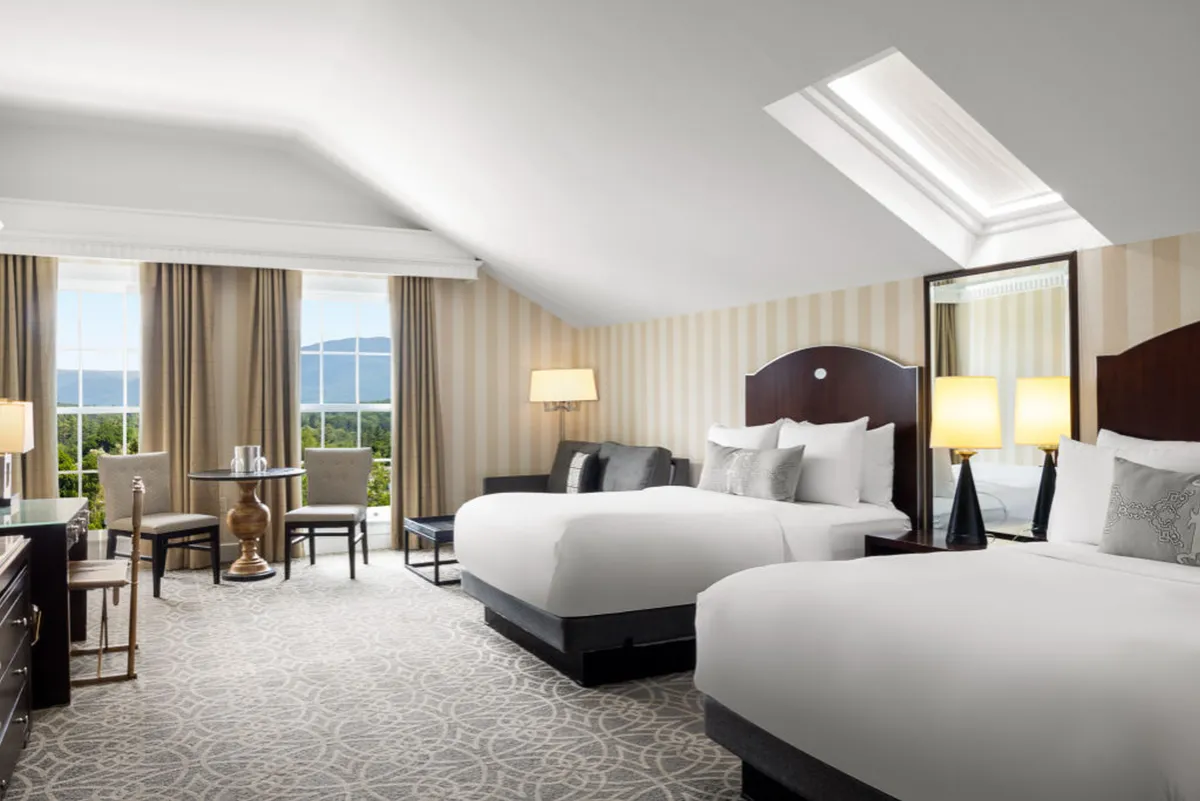 The image features a spacious, inviting studio with two plush queen beds adorned with decorative pillows, a cozy armchair, and a wooden desk and chairs. A large window offers breathtaking views of the Vermont mountains, filling the room with natural light and welcoming the tranquil beauty of the outdoors inside.