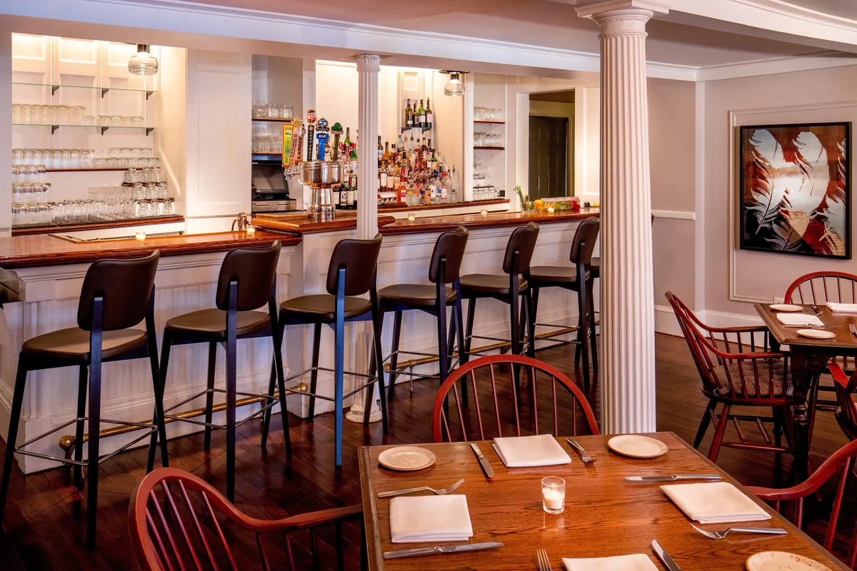 This image showcases a stylish interior bar area with high stools, carefully set tables, and contemporary artwork adorning the walls. The ambiance exudes casual sophistication and comfort, making it an inviting space for guests to relax and socialize.